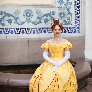 May include: A woman in a yellow gown with red trim sits on a stone bench. She is wearing white gloves and a pearl necklace. The background is a tiled wall with blue and white floral designs.
