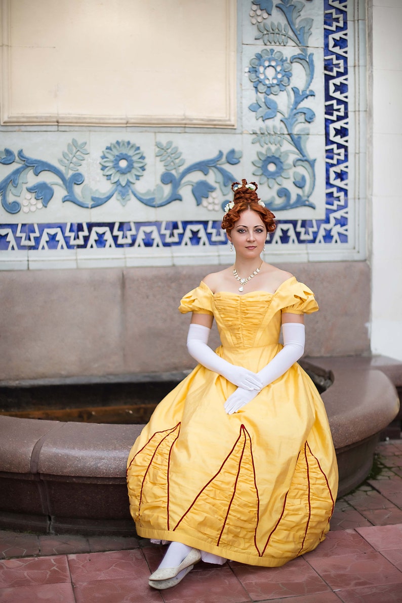 May include: A woman in a yellow gown with red trim sits on a stone bench. She is wearing white gloves and a pearl necklace. The background is a tiled wall with blue and white floral designs.