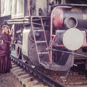 Op de afbeelding: Een vrouw in een bordeauxrode jurk staat voor een grote, vintage stoomlocomotief. De trein staat op rails en heeft een grote, ronde koplamp. De vrouw houdt zich vast aan een leuning aan de zijkant van de trein.