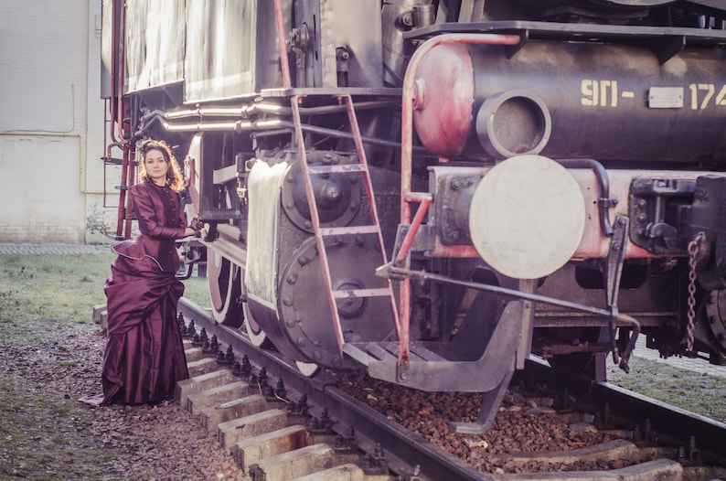 Op de afbeelding: Een vrouw in een bordeauxrode jurk staat voor een grote, vintage stoomlocomotief. De trein staat op rails en heeft een grote, ronde koplamp. De vrouw houdt zich vast aan een leuning aan de zijkant van de trein.