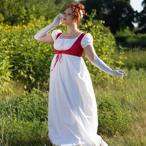 May include: A white Regency-style dress with a red bodice and white gloves. The dress features a square neckline and short, puffed sleeves. A pearl necklace and a flower in the hair complete the ensemble. The dress is worn outdoors in a field of green.