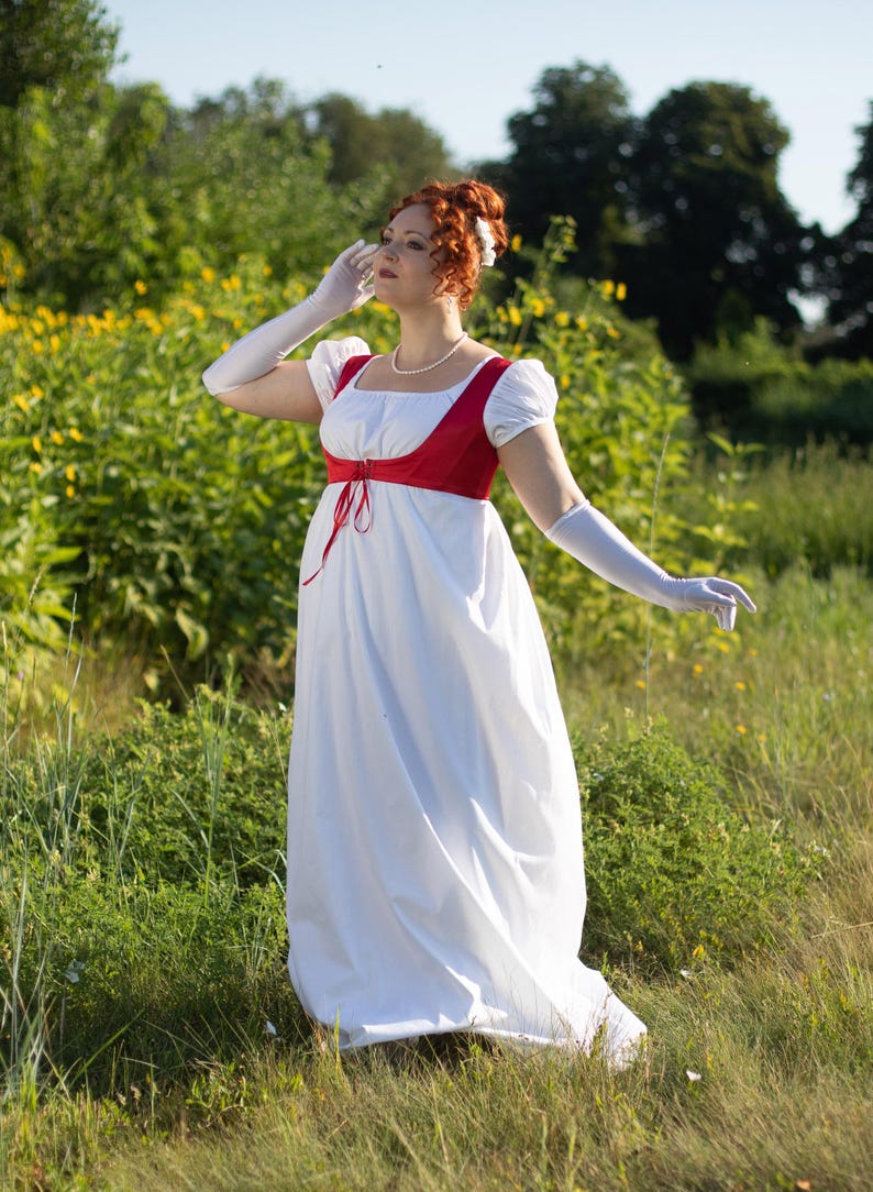May include: A white Regency-style dress with a red bodice and white gloves. The dress features a square neckline and short, puffed sleeves. A pearl necklace and a flower in the hair complete the ensemble. The dress is worn outdoors in a field of green.