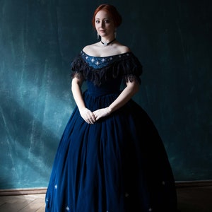May include: A woman wearing a blue velvet gown with a black lace bodice and white star embellishments. She is standing in front of a dark green background.