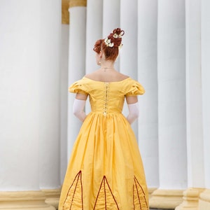 May include: A woman in a yellow dress with a white lace bodice and a red trim. The dress has a full skirt with a ruffled hem. She is standing in front of a white column.