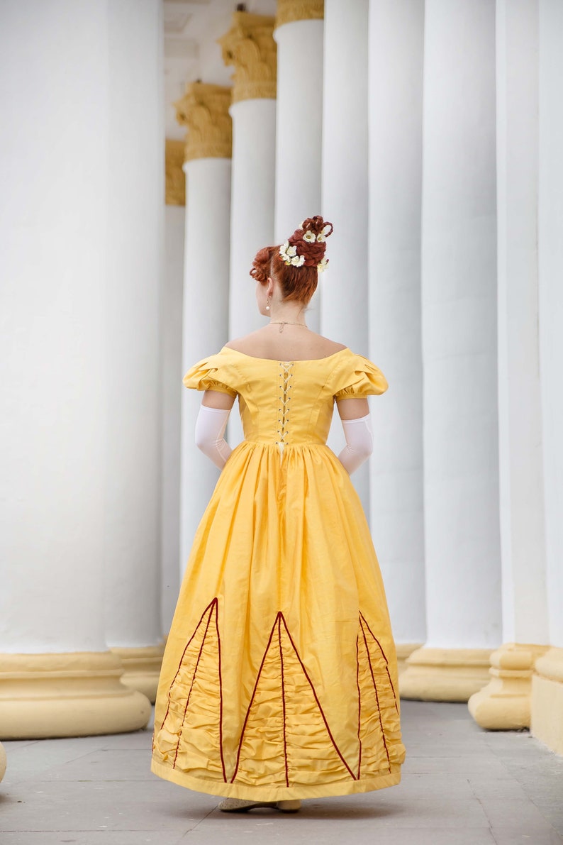 May include: A woman in a yellow dress with a white lace bodice and a red trim. The dress has a full skirt with a ruffled hem. She is standing in front of a white column.