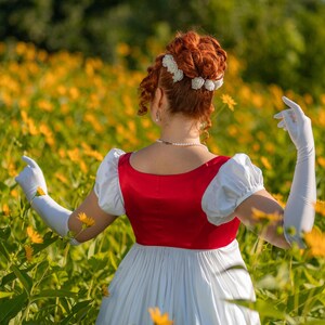 May include: A person in a red and white dress with long white gloves stands in a field of yellow flowers. The dress has a red bodice and a white skirt. The person's hair is styled in an updo with white flower accents.