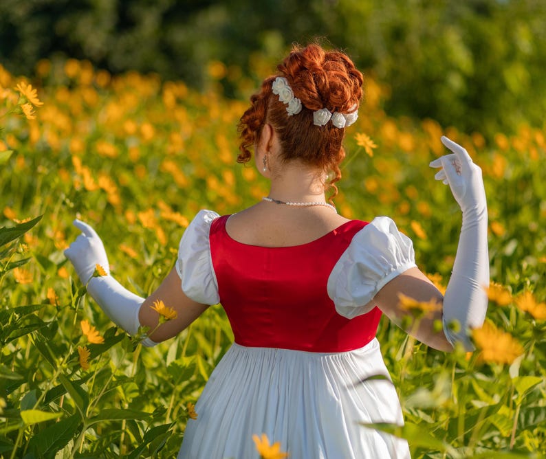May include: A person in a red and white dress with long white gloves stands in a field of yellow flowers. The dress has a red bodice and a white skirt. The person's hair is styled in an updo with white flower accents.