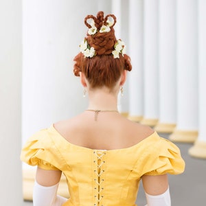 May include: A woman wearing a yellow dress with a lace-up back and white gloves. Her hair is styled in a high updo with white flowers.