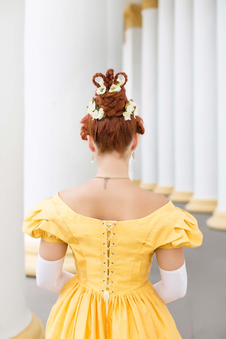 May include: A woman wearing a yellow dress with a lace-up back and white gloves. Her hair is styled in a high updo with white flowers.