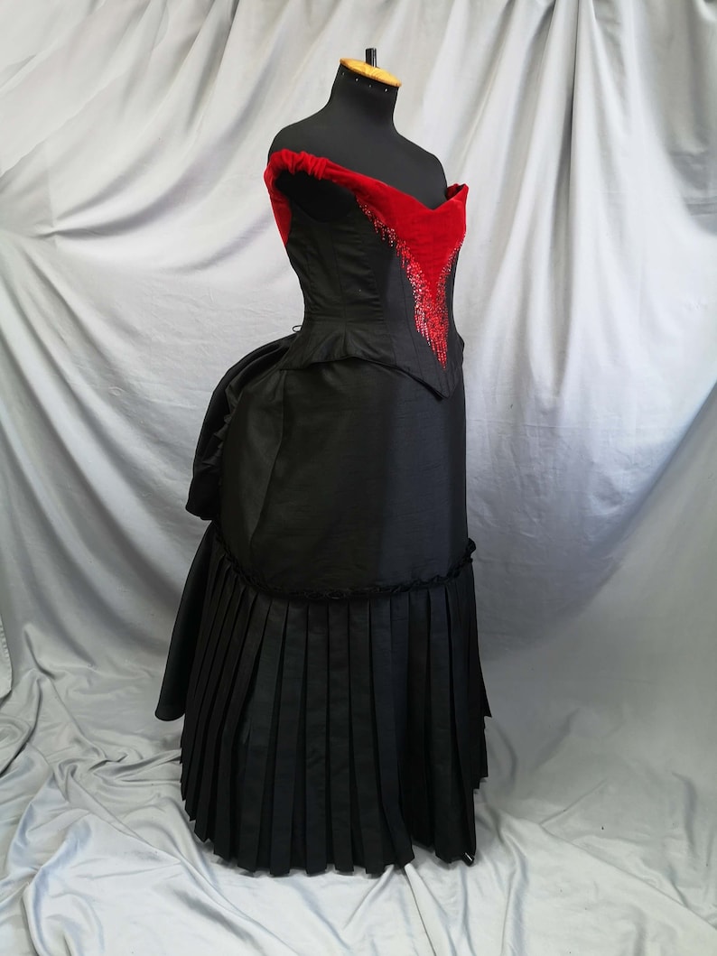 Op de afbeelding: Een zwarte en rode galajurk met een sweetheart-halslijn en een strak lijfje. Het lijfje is versierd met rood fluweel en een parelmoerontwerp. De rok is vol en zwierig met plooien.