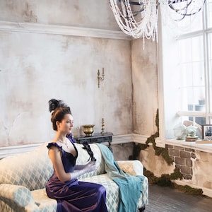 May include: A woman in a purple dress sits on a patterned couch in a room with a chandelier and a window. She is holding a black feather fan.