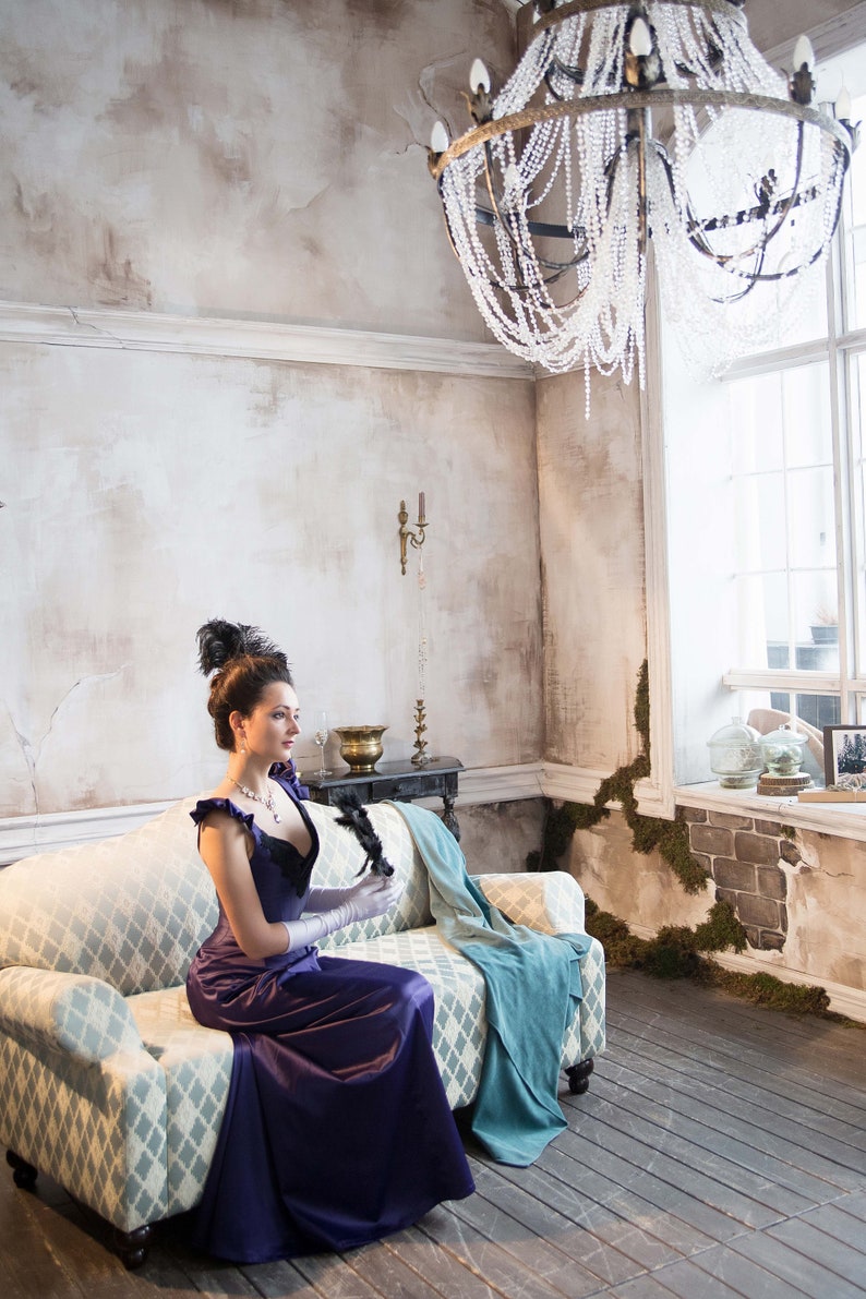 May include: A woman in a purple dress sits on a patterned couch in a room with a chandelier and a window. She is holding a black feather fan.