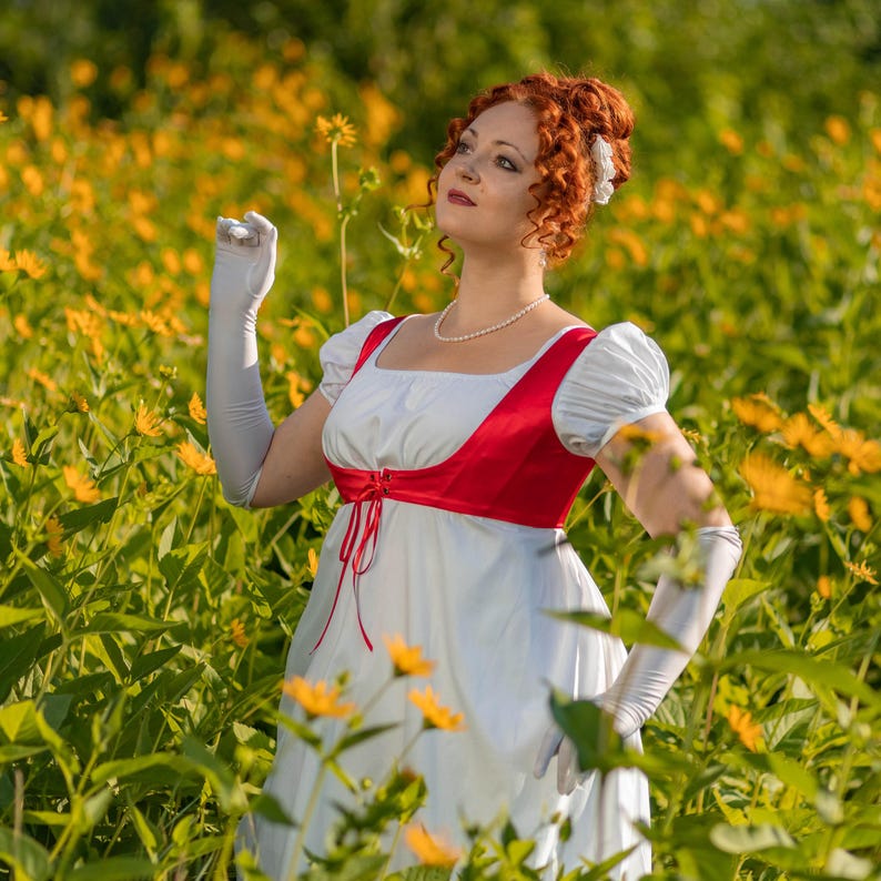 May include: A woman in a white Regency-style dress with a red bodice and white gloves stands in a field of yellow flowers. The dress features a square neckline and short, puffed sleeves. A pearl necklace and red hair complete the look.