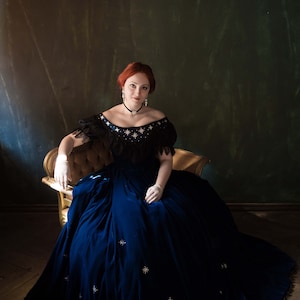 May include: A woman in a blue velvet gown with a black lace bodice and white gloves sits on a gold chair against a dark green background. The gown is adorned with silver stars.