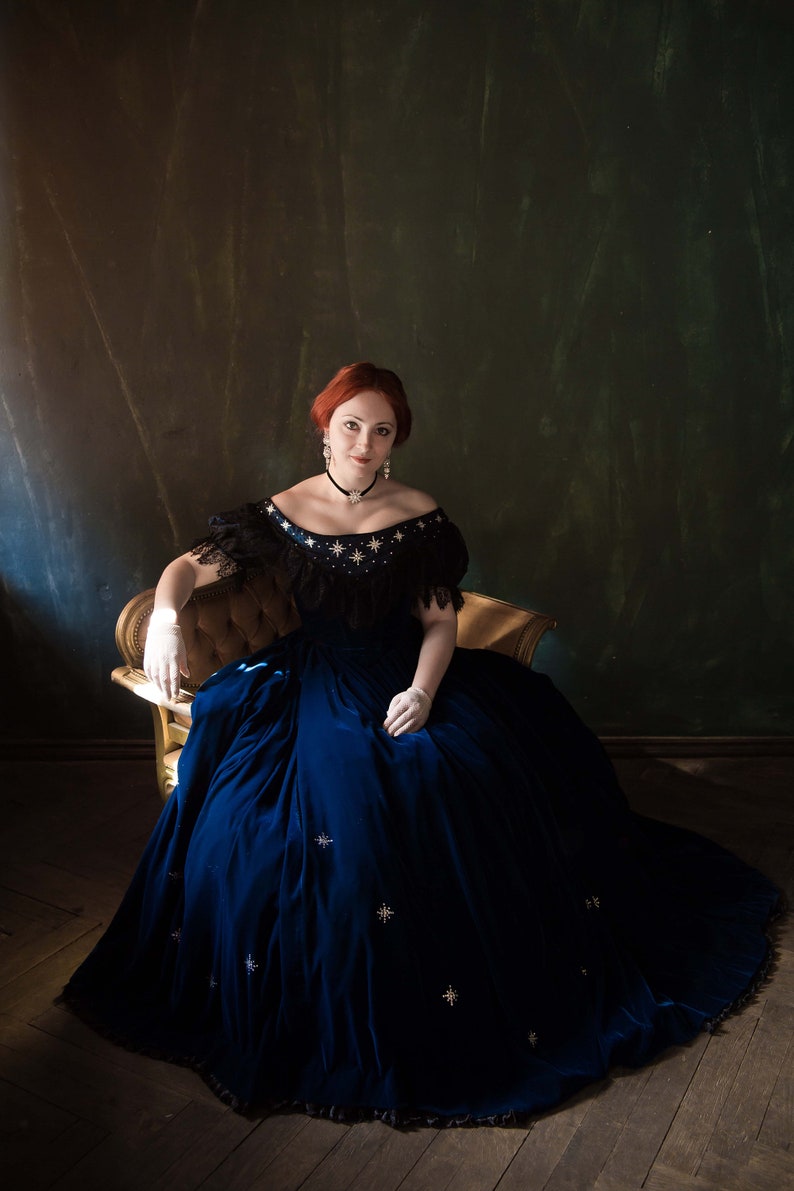 May include: A woman in a blue velvet gown with a black lace bodice and white gloves sits on a gold chair against a dark green background. The gown is adorned with silver stars.