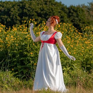 May include: A white Regency-style dress with a red bodice and short sleeves. The dress features a high waist and a flowing skirt. The model is wearing white gloves and a pearl necklace. The background is a field of yellow flowers.