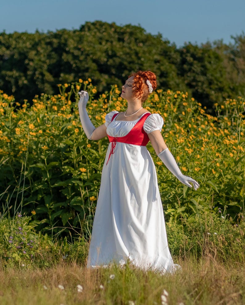 May include: A white Regency-style dress with a red bodice and short sleeves. The dress features a high waist and a flowing skirt. The model is wearing white gloves and a pearl necklace. The background is a field of yellow flowers.