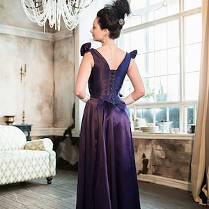 May include: A woman in a purple satin dress with a lace-up back stands in a room with a chandelier and a window.