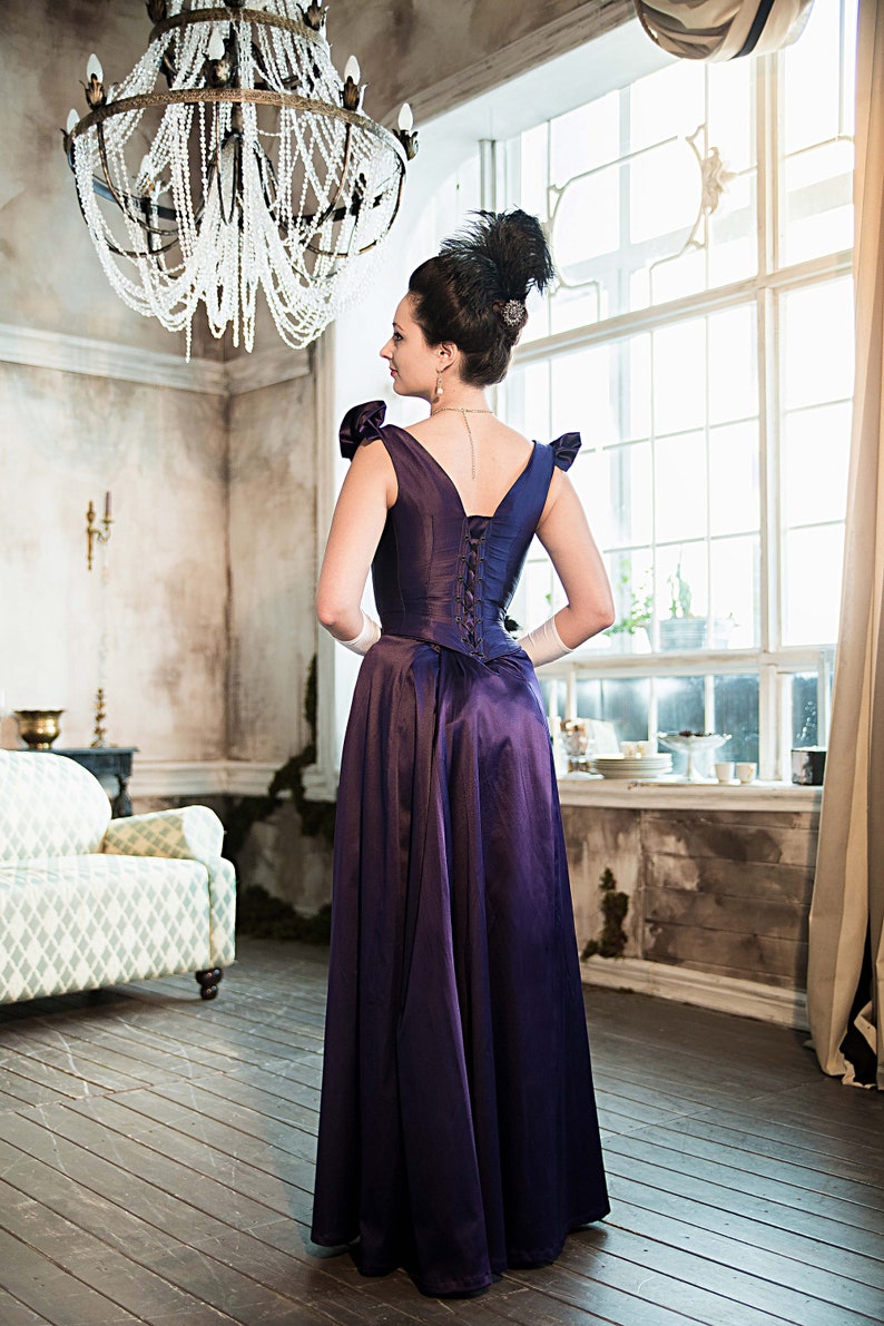 May include: A woman in a purple satin dress with a lace-up back stands in a room with a chandelier and a window.