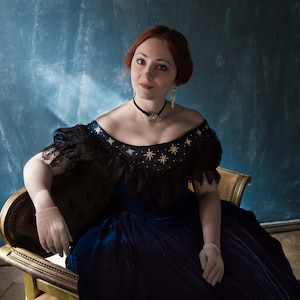 May include: A woman with red hair wearing a blue velvet dress with black lace trim and gold star embellishments. She is sitting on a gold chair against a blue background.