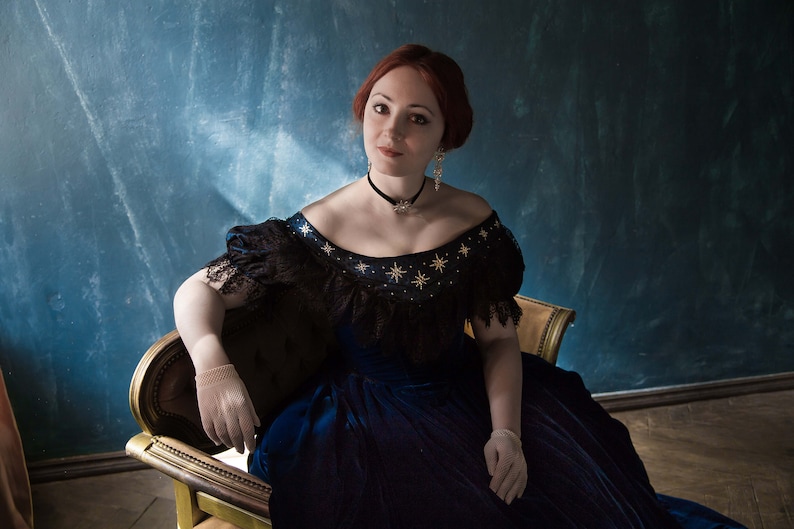May include: A woman with red hair wearing a blue velvet dress with black lace trim and gold star embellishments. She is sitting on a gold chair against a blue background.