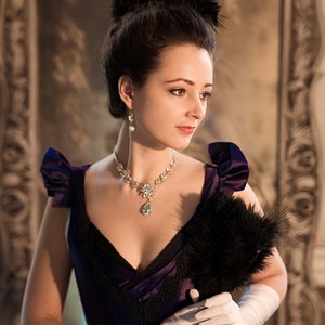 May include: A woman in a purple dress with a black feather boa and white gloves. She is wearing a pearl necklace and earrings.