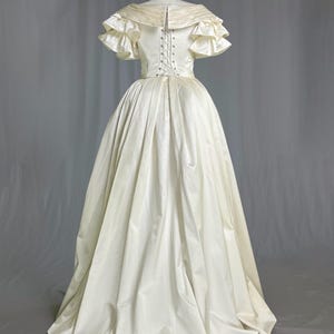 May include: A white satin wedding dress with a fitted bodice and a full skirt. The dress has a lace-up back and short, ruffled sleeves.