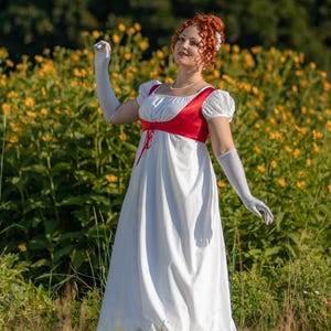 May include: A white empire waist dress with a red bodice and white sleeves. The dress features a high waistline and a flowing skirt. The sleeves are short and puffed. The person is wearing long white gloves and a pearl necklace.