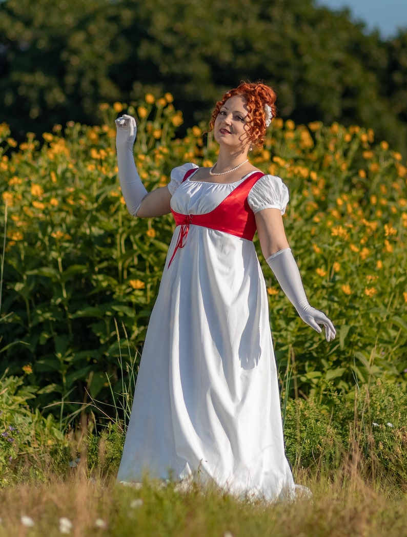 May include: A white empire waist dress with a red bodice and white sleeves. The dress features a high waistline and a flowing skirt. The sleeves are short and puffed. The person is wearing long white gloves and a pearl necklace.