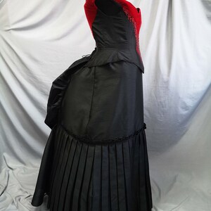 Op de afbeelding: Een zwarte en rode galajurk met een strak lijfje en een volle rok. Het lijfje heeft een rode fluwelen overlay en is versierd met pailletten. De rok is geplooid en heeft een bustle aan de achterkant.
