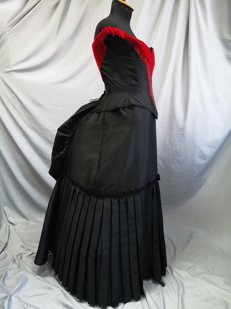Op de afbeelding: Een zwarte en rode galajurk met een strak lijfje en een volle rok. Het lijfje heeft een rode fluwelen overlay en is versierd met pailletten. De rok is geplooid en heeft een bustle aan de achterkant.