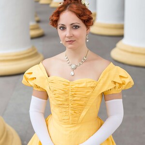 May include: A woman in a yellow gown with a white lace collar and white gloves. She has her hair up in a bun with white flowers. She is standing in front of a white column.