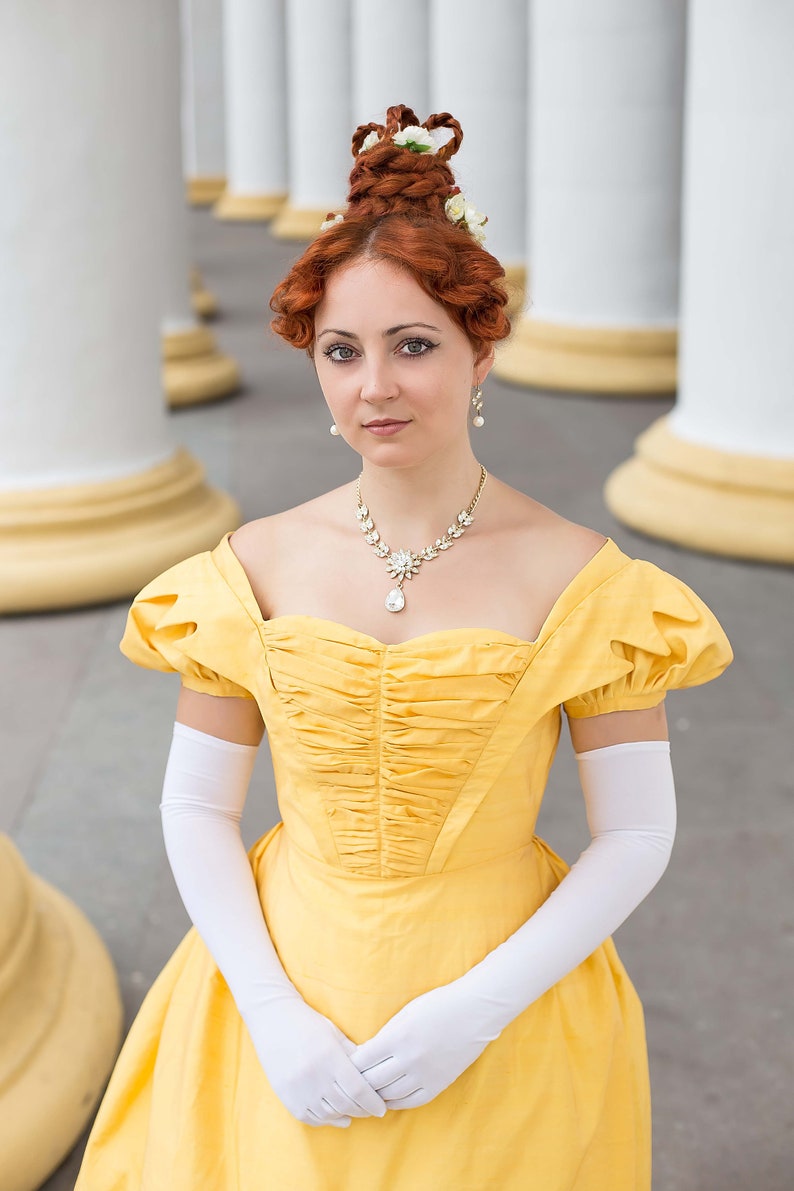 May include: A woman in a yellow gown with a white lace collar and white gloves. She has her hair up in a bun with white flowers. She is standing in front of a white column.