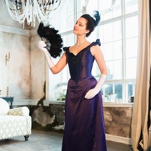 May include: A woman in a purple dress with a black lace bodice and a black feather in her hair holds a black feather fan in her right hand. She is wearing white gloves and a pearl necklace.