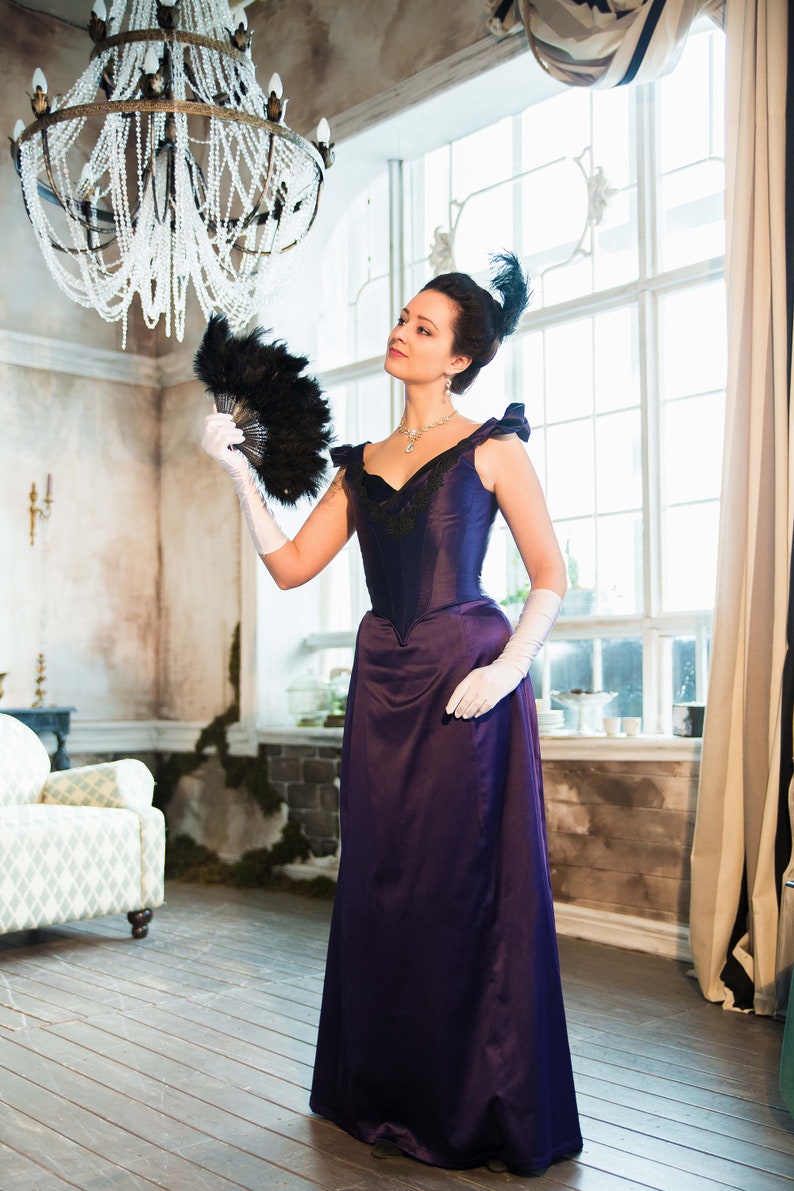 May include: A woman in a purple dress with a black lace bodice and a black feather in her hair holds a black feather fan in her right hand. She is wearing white gloves and a pearl necklace.