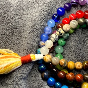 Könnte beinhalten: Eine bunte Perlenkette mit einem Quasten aus gelbem und orangefarbenem Garn. Die Perlen bestehen aus verschiedenen Steinen und Mineralien, darunter Amethyst, Achat und Jaspis.