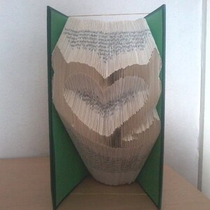 May include: A folded book art piece with a heart shape carved into the pages. The book is open, revealing the heart design, and is framed by green covers. The pages are beige, and the overall effect is a unique decorative item.