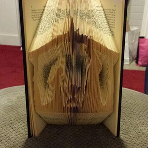 May include: A book sculpture with pages folded to create a three-dimensional architectural design. The book is open, revealing the intricate folded pages, with a dark cover and a light-colored interior.