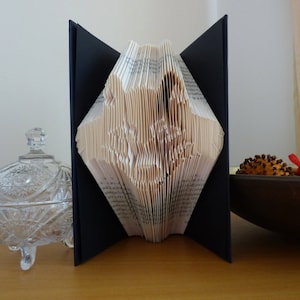 May include: A decorative book with pages folded into a diamond shape. The book is open, revealing the intricate design of the folded pages. The book is set against a neutral background, with a glass container and wooden bowl in the foreground.