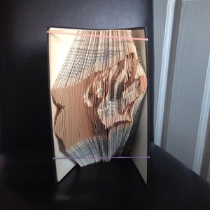 May include: A book with pages folded to create a three-dimensional design. The folded pages form a shape, possibly a star or a similar geometric figure. The book is held together with pink and purple rubber bands.