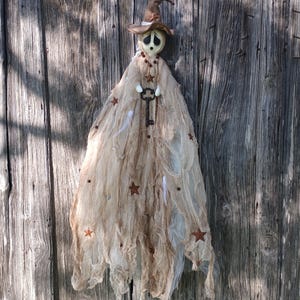 May include: A hanging ghost decoration featuring a pale, round face with large black eye sockets, wearing a brown pointed hat. The figure is draped in layers of tattered, beige cheesecloth fabric adorned with small brown stars and a metal key.