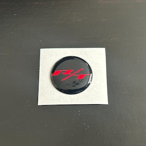 May include: A black, circular emblem with the red letters "R/T" is centered on a square, white sticker. The emblem has a glossy finish and is likely a car accessory.