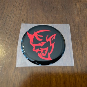 May include: A red and black round sticker with a stylized red devil head graphic. The sticker is on a clear backing.