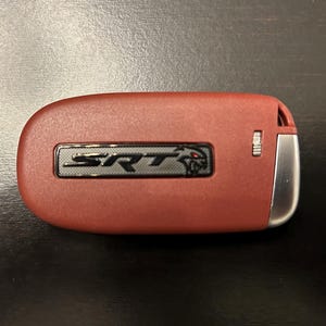 May include: A red car key fob with the letters "SRT" in black, and a silver accent. The fob has a textured surface and a small, red devil logo.