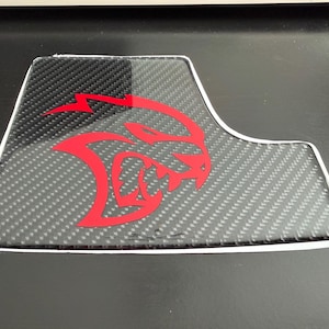 May include: A carbon fiber-style car accessory with a red graphic of a stylized animal head and lightning bolt. The accessory has a white border and is set against a dark background.