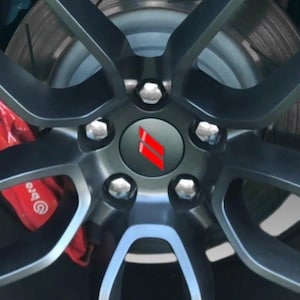 May include: Close-up of a car wheel with a dark gray finish. The wheel features a central hubcap with a red and white logo. The brake calipers are red, and the wheel is secured with silver lug nuts.