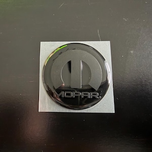 May include: A glossy black Mopar emblem with the Mopar logo and the word "MOPAR" in a sans-serif font. The emblem is circular and set on a square, silver backing.
