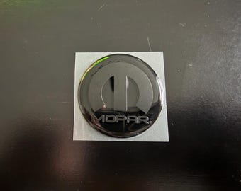 Insignia MOPAR para la tapa del depósito de combustible del Challenger. M y Mopar en negro mate. Fondo negro brillante.