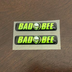 May include: Two black rectangular stickers with the words "BAD BEE" in neon green, featuring a cartoon bee wearing a helmet. The stickers are on a silver backing, set against a brown wooden surface.