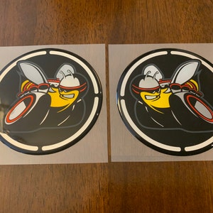 May include: Two black and white round stickers with a white border. The stickers feature a cartoon bee with yellow and black stripes, red eyes, and a black helmet. The bee is driving a car with red and gray wheels.